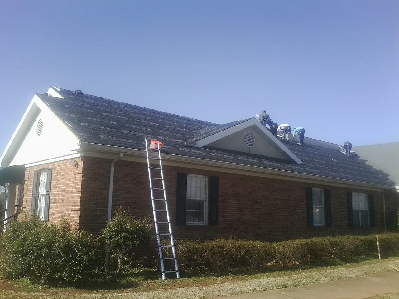 Crew tearing shingles off a brick ranch roof with a ladder set — Emergency Roof Repair work by our crew in Nesconset