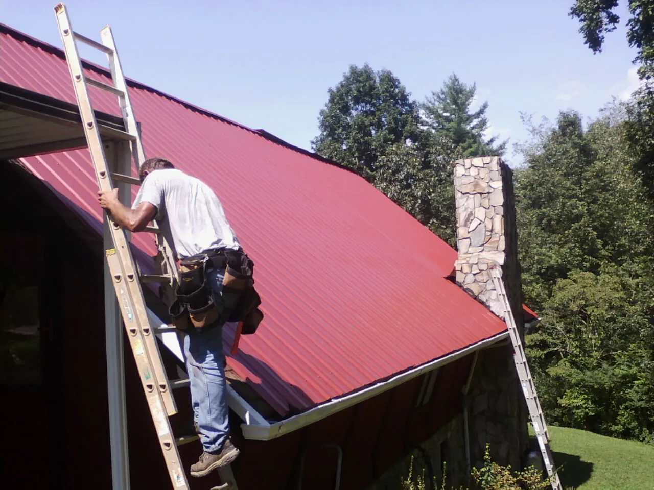 Roofer on a ladder beside a red metal roof and stone chimney — Soffit & Fascia Repair by Roofing Brothers in Nesconset